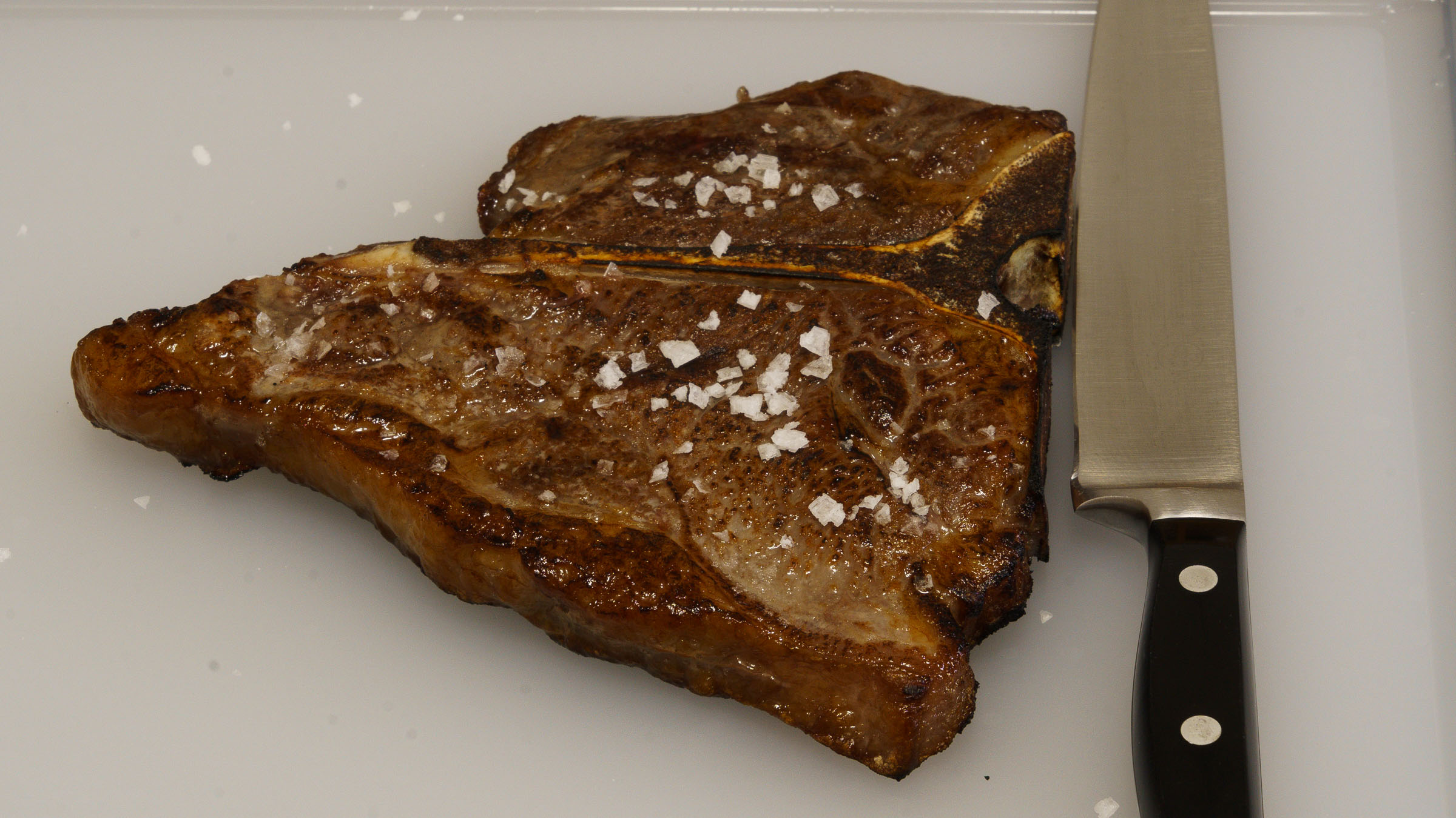Cooked T-bone steak with flaky salt and a large knife.
