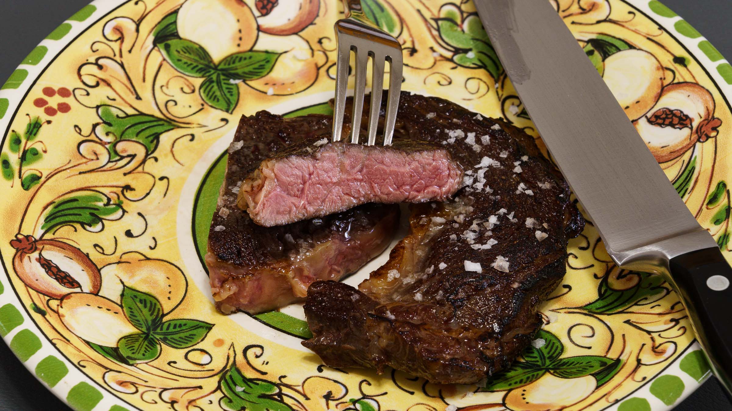 A brightly coloured dinner plate with a steak. The photograph focusses on a piece of steak being held by a fork to demonstrate the perfect edge-to-edge pinkness. The meat has been seasoned with flaky salt. My best steak knife is also in the photograph.