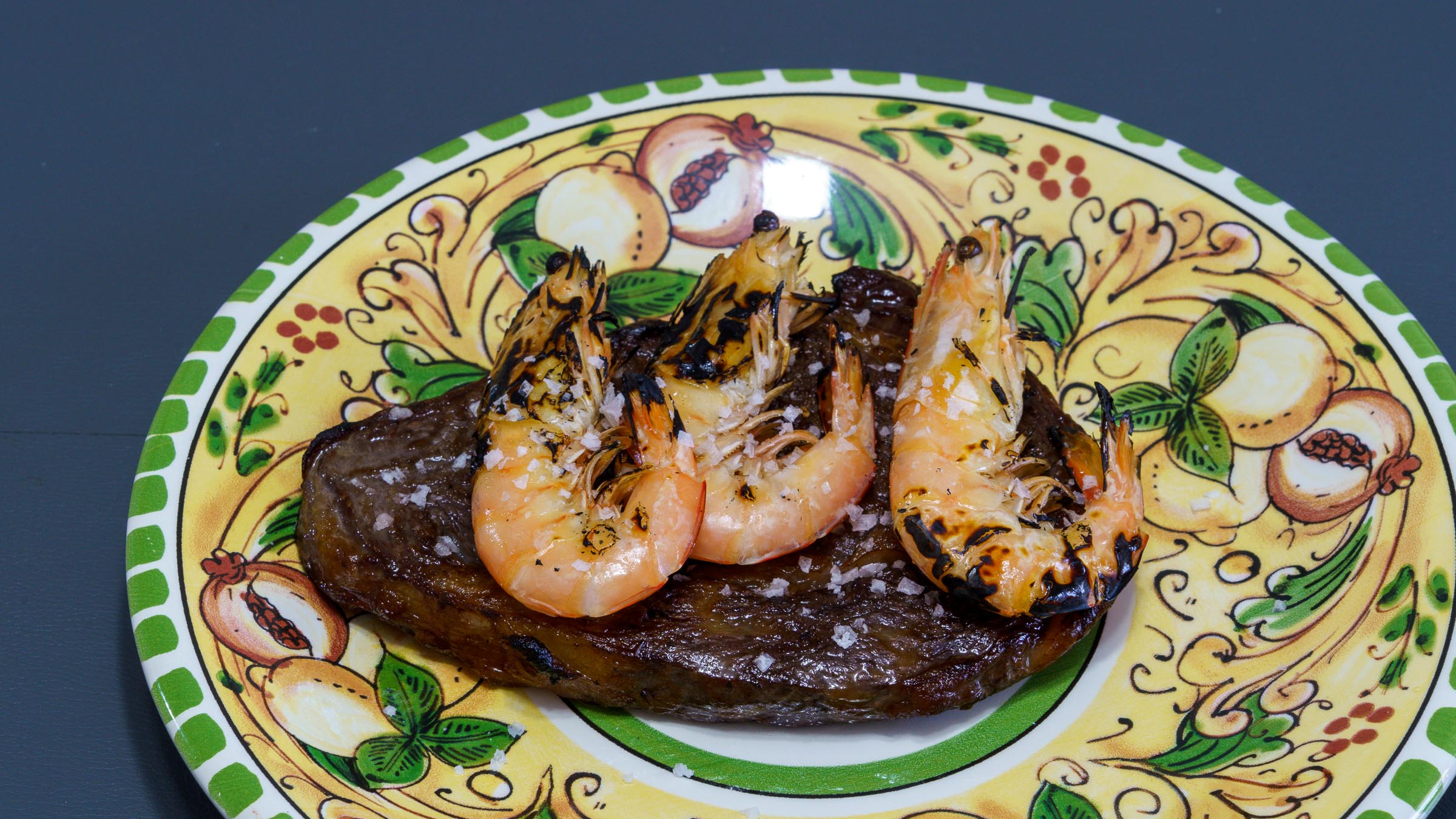 A floral dinner plate featuring pomegranates with a perfectly cooked scotch fillet steak and three freshly flame torched South Australian king prawns