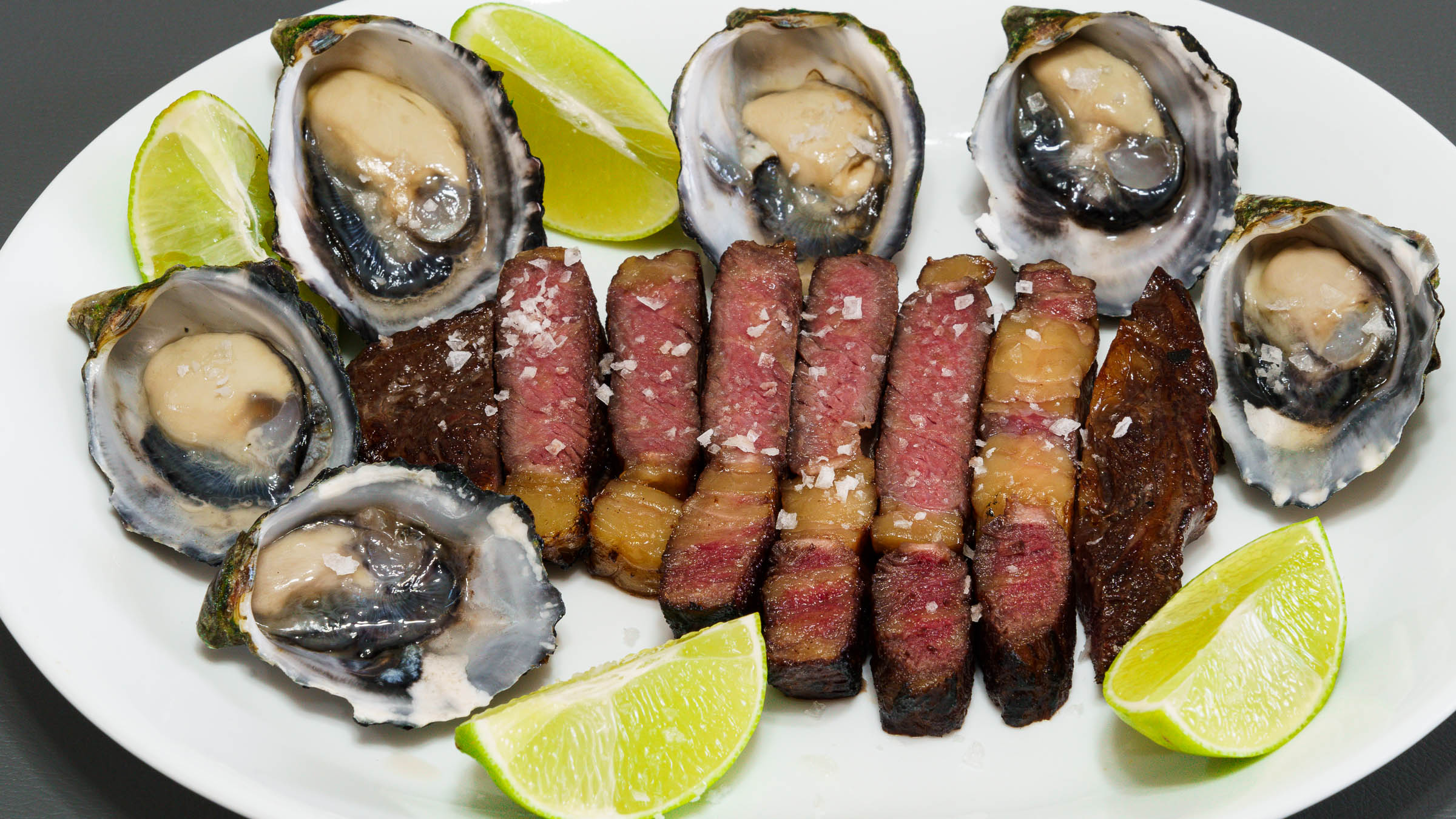 Dinner plate with sliced scotch fillet steak and six oysters in their shells with lime wedges and salt. The focus is on the far left oyster.