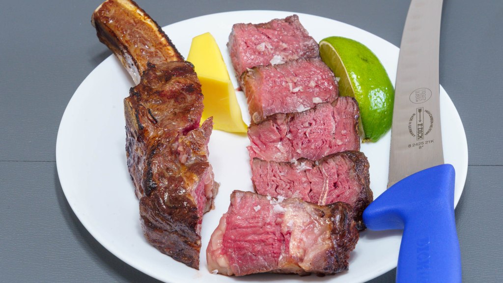 A photograph of a bone-in ribeye steak with the bone and sliced deckle (spinalis) meat. Also in the photograph is a wedge of lime, a wodge of butter, and a butchers knife.