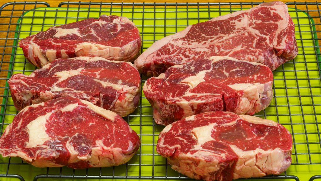 A photograph of a refrigerator rack with six steak on it. One is a Wagyu porterhouse steak in the top right corner. The other five steaks are economy-grade scotch fillet steak, each slice is about 3 cm thick.