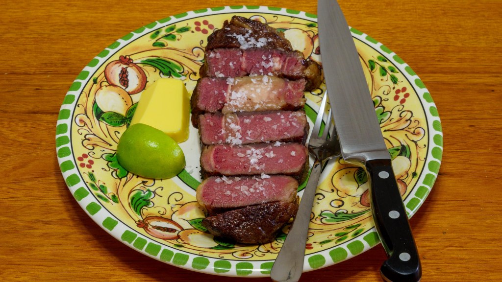 A photograph of a piece of scotch fillet steak sliced thickly and seasoned with flaky salt. The dinner plate is floral with pomegranate halves. There is a wedge of lime and a bit of butter on the plate plus a carving knife and a fork.