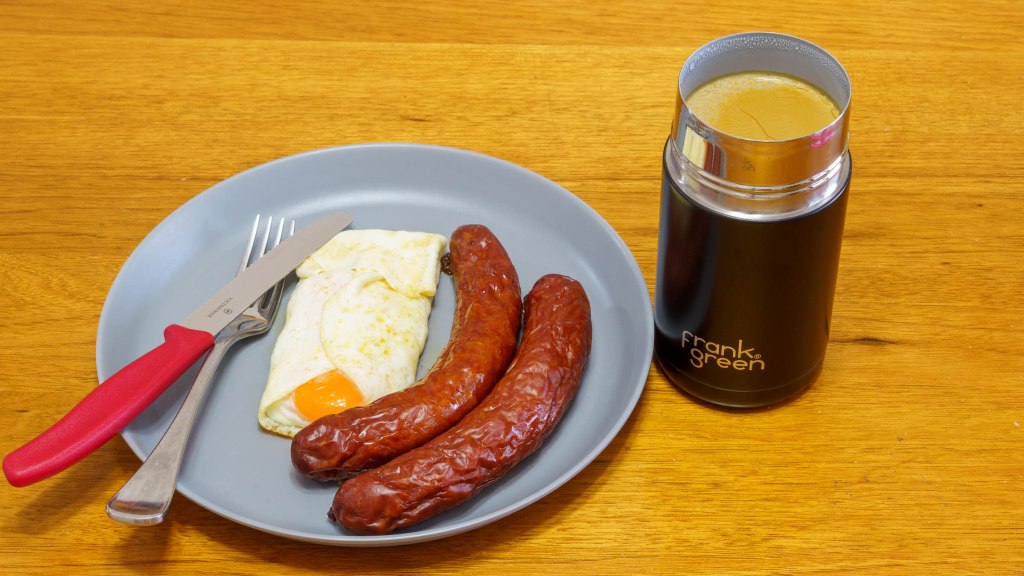 A photograph of a grey plate with two cheese kransky sausages and two eggs fried in butter. The photograph also had a new red handled Victorinox steak knife and a black frank green cup containing coffee made with cream (which is splitting)