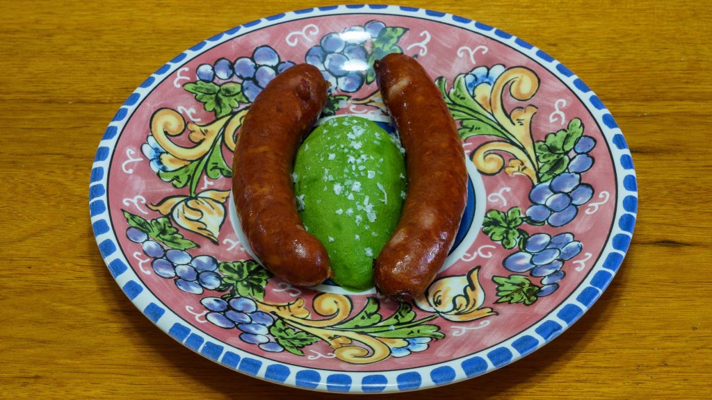 A photograph of two cheese kransky sausages arranged either side of a cheek of Shepard avocado. The avocado is seasoned with flaky salt.