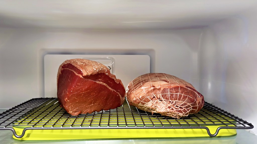 A photograph of the top shelf of my refrigerator with a piece of beef and a piece of lamb on a rack.