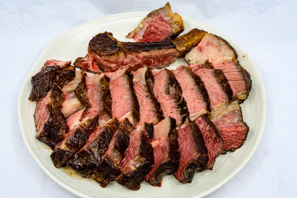 Photograph of Bone-in ribeye steak after being seared on a hot cast-iron flat top barbecue grill with a gas torch. Dissected from the rib bone and the spinalis separated from the longissimus dorsi muscle. Both muscle groups have been sliced and laid out on the dinner plate.