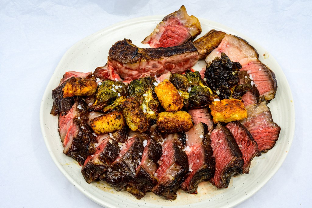 Photograph of Bone-in ribeye steak after being seared on a hot cast-iron flat top barbecue grill with a gas torch. Dissected from the rib bone and the spinalis separated from the longissimus dorsi muscle. Both muscle groups have been sliced and laid out on the dinner plate. The potato gems and Brussels sprouts have been added on top of the cut meat.