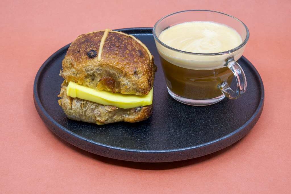 A freshly toasted sourdough Hot Cross bun from Perryman’s Bakery in North Adelaide is shown. A large wedge of grass-fed butter is placed between the top and bottom of the bun. Next to the bun on the right is a clear glass cup of coffee with a lovely crema on top. The bun and coffee are placed on a black ceramic plate, which is sitting on butcher’s paper.