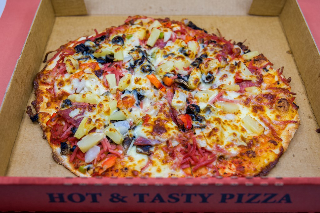 A box of Oasis Pizzeria’s 'Hot and Tasty Pizza' is shown. The pizza is topped with black olives, pepperoni, pineapple, bacon, mozzarella cheese, and anchovies.