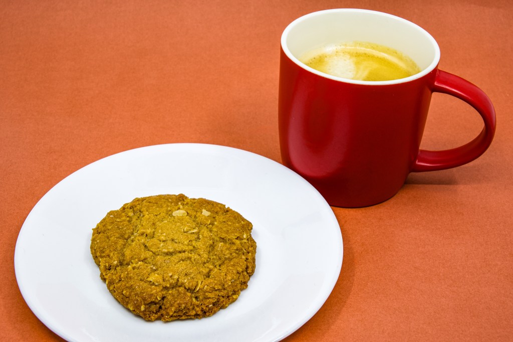 A photograph of a Anzac biscuit on a white saucer and a mug coffee in a red mug