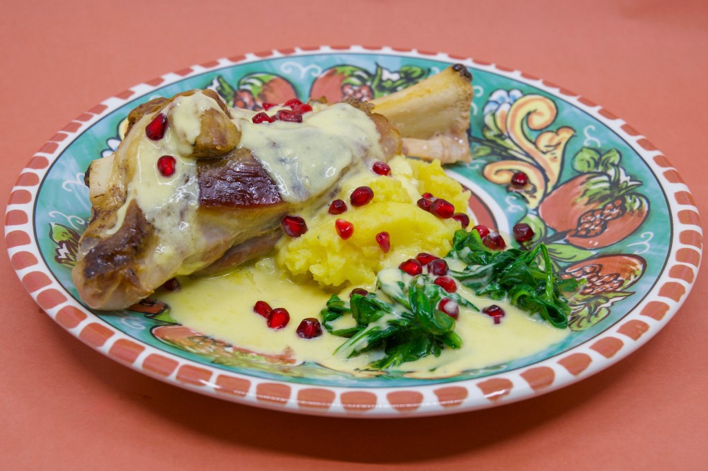 Slow-cooked lamb shank on potato mash with wilted spinach leaves, served with a blue-veined cheese sauce and garnished with pomegranate arils. On a dinner plate featuring pomegranates.