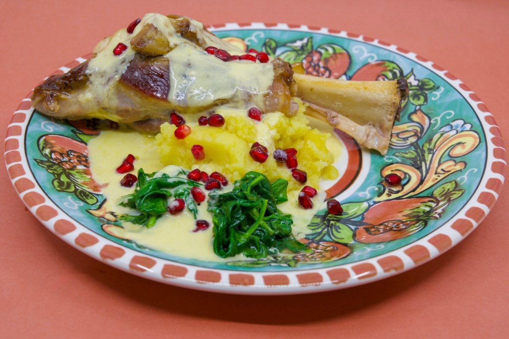 Slow-cooked lamb shank on potato mash with wilted spinach leaves, served with a blue-veined cheese sauce and garnished with pomegranate arils. On a dinner plate featuring pomegranates.