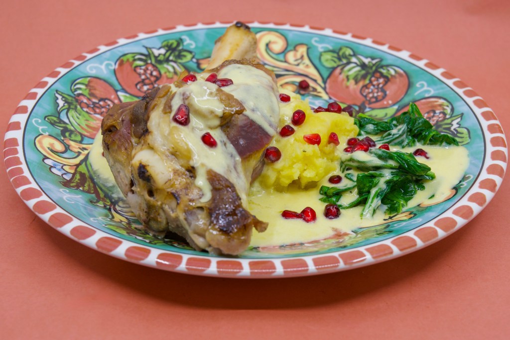 Slow-cooked lamb shank on potato mash with wilted spinach leaves, served with a blue-veined cheese sauce and garnished with pomegranate arils. On a dinner plate featuring pomegranates.