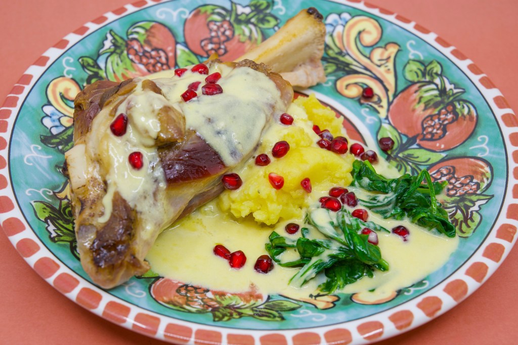 Slow-cooked lamb shank on potato mash with wilted spinach leaves, served with a blue-veined cheese sauce and garnished with pomegranate arils. On a dinner plate featuring pomegranates.