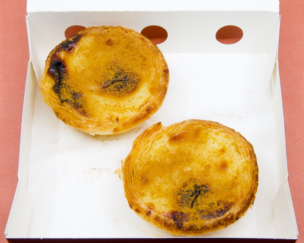 A photograph of two Portuguese custard tarts dusted with cinnamon powder from Saudade Portuguese Tarts in Adelaide Central Market.