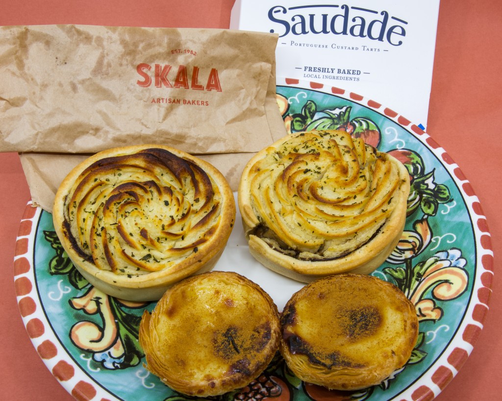 A photograph of a colourful plate with two potato pies and two Portuguese tarts. There is also a brown paper bag from Skala Artisan Bakeries plus the cardboard top for Saudade Portuguese Custard Tarts.
