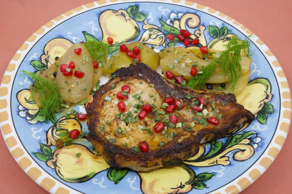A photograph of a colourful (warmed) plate with a grilled pork chop and stewed fennel and apple. The meal is garnished with fennel fronds and pickled pomegranate arils.