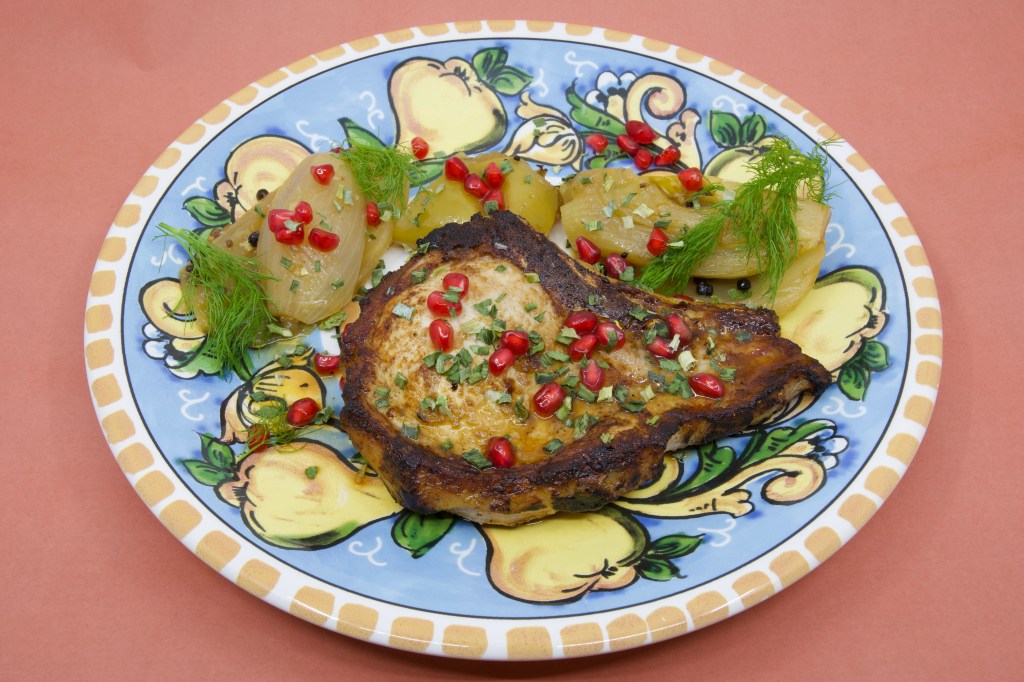 A photograph of a colourful (warmed) plate with a grilled pork chop and stewed fennel and apple. The meal is garnished with fennel fronds and pickled pomegranate arils.