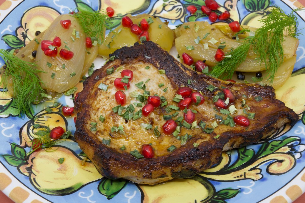 A photograph of a colourful (warmed) plate with a grilled pork chop and stewed fennel and apple. The meal is garnished with fennel fronds and pickled pomegranate arils.