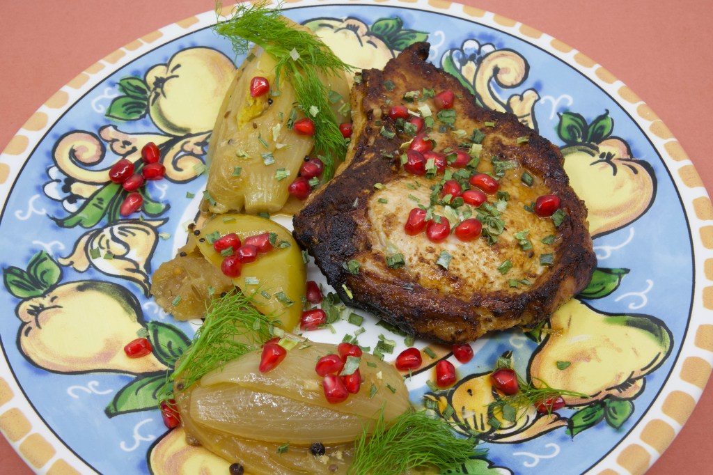 A photograph of a colourful (warmed) plate with a grilled pork chop and stewed fennel and apple. The meal is garnished with fennel fronds and pickled pomegranate arils.