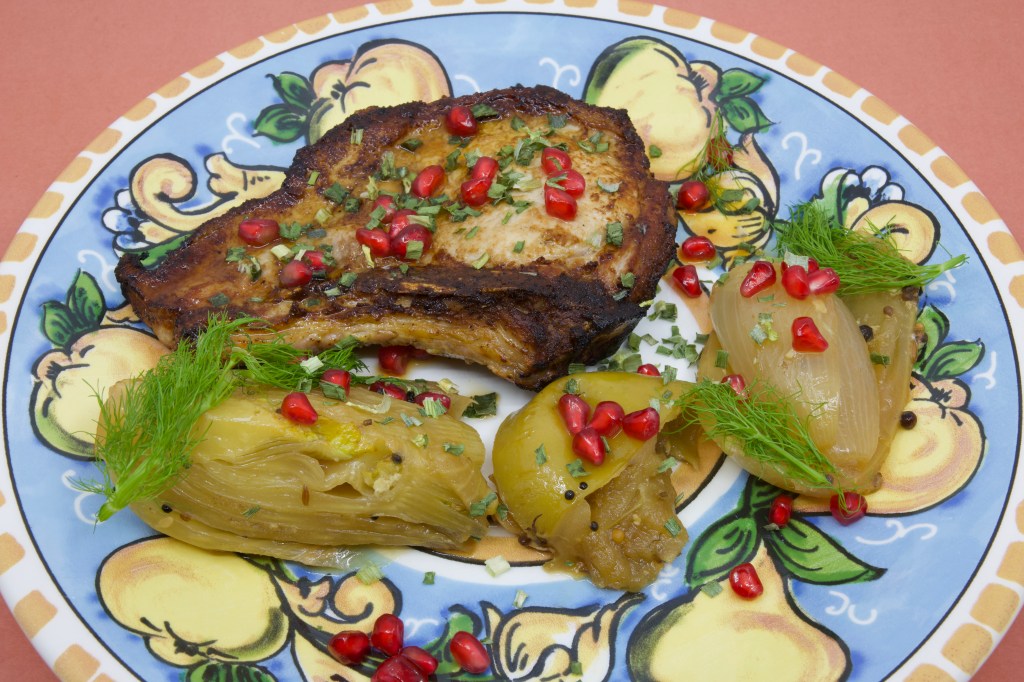 A photograph of a colourful (warmed) plate with a grilled pork chop and stewed fennel and apple. The meal is garnished with fennel fronds and pickled pomegranate arils.