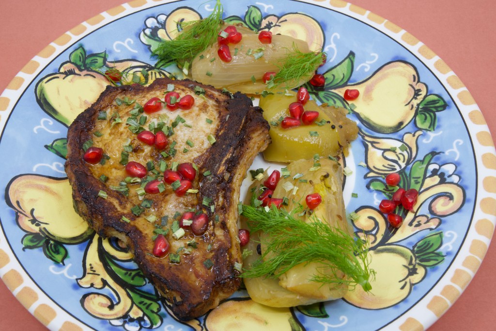 A photograph of a colourful (warmed) plate with a grilled pork chop and stewed fennel and apple. The meal is garnished with fennel fronds and pickled pomegranate arils.