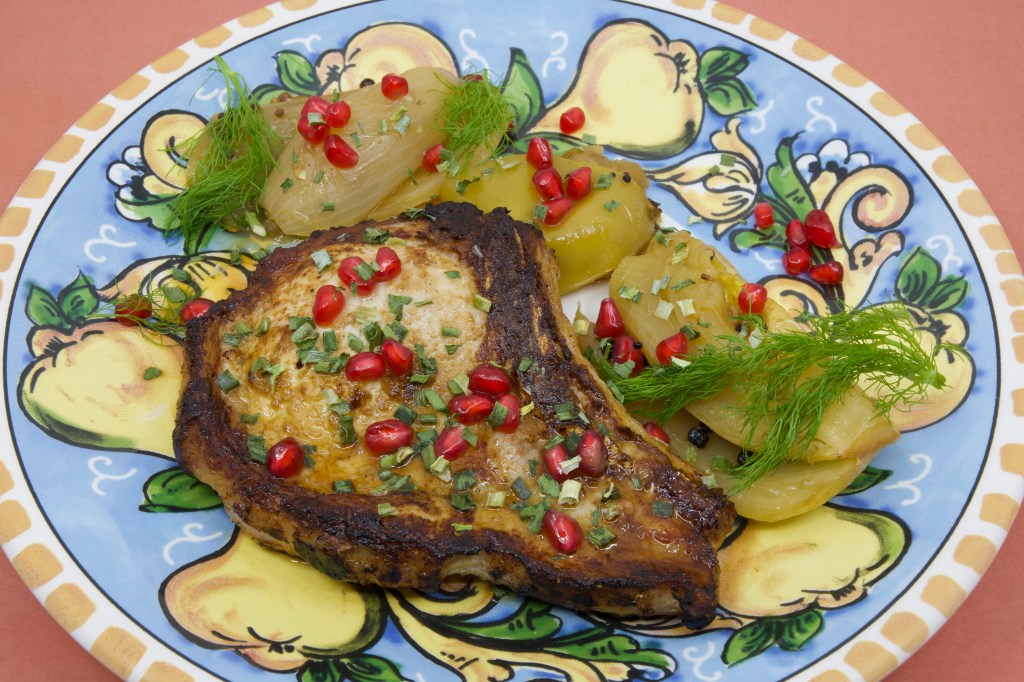 A photograph of a colourful (warmed) plate with a grilled pork chop and stewed fennel and apple. The meal is garnished with fennel fronds and pickled pomegranate arils.