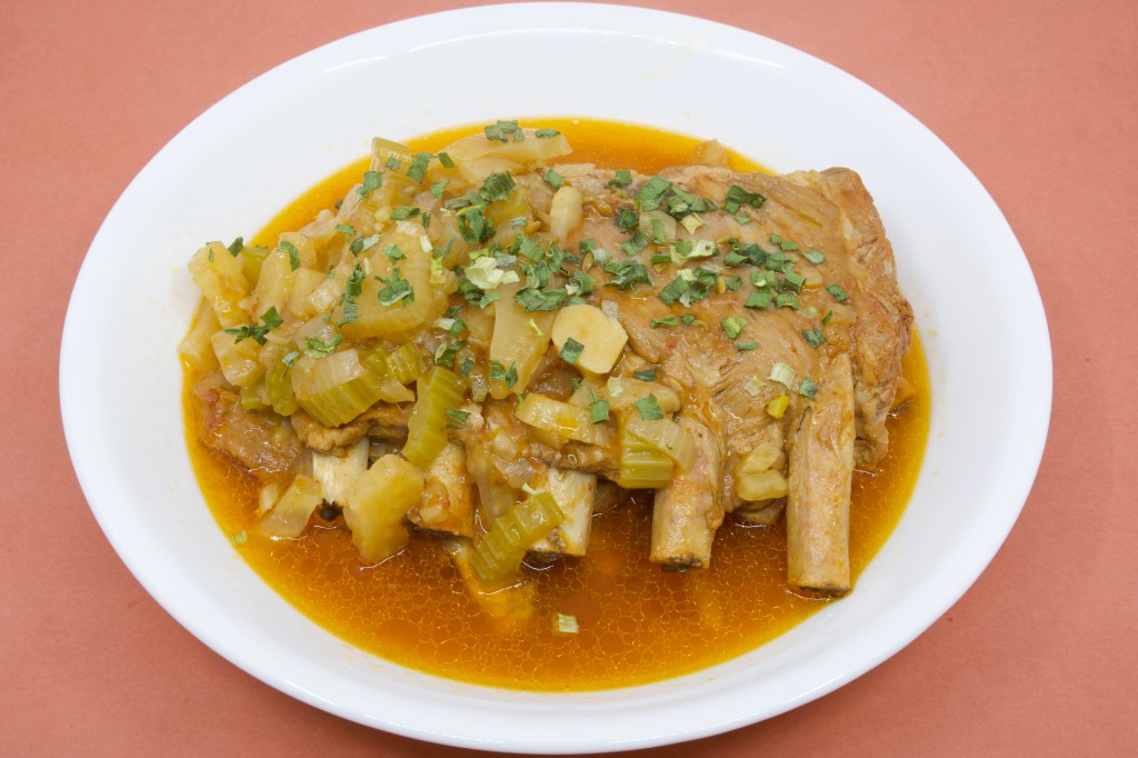 A photograph of a shallow white ceramic bowl containing two racks of pork ribs that had been cooked in the pressure cooker. The ribs are sitting in a thin broth of the cooking liquor, along with soft fennel, onion, and celery. The dish is garnished with some chives.