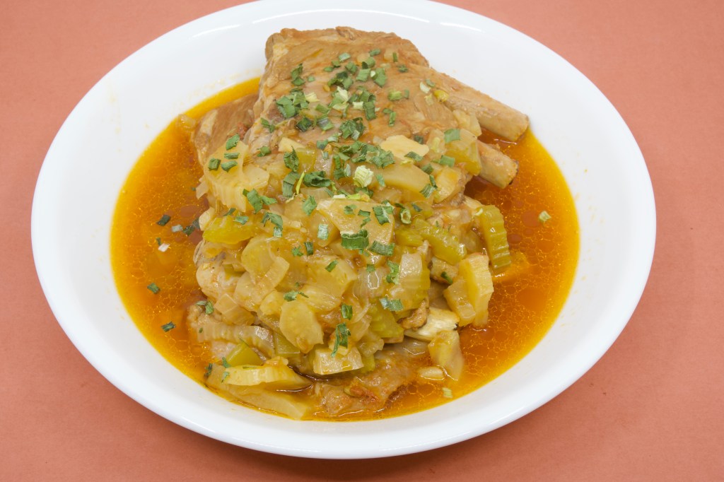A photograph of a shallow white ceramic bowl containing two racks of pork ribs that had been cooked in the pressure cooker. The ribs are sitting in a thin broth of the cooking liquor, along with soft fennel, onion, and celery. The dish is garnished with some chives.