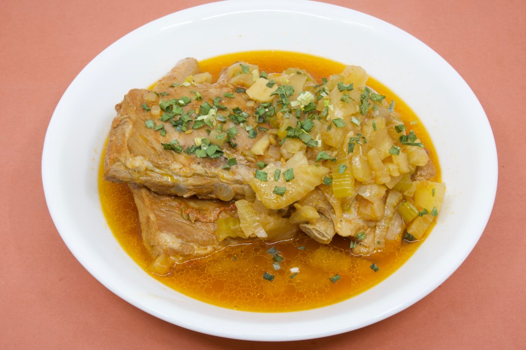 A photograph of a shallow white ceramic bowl containing two racks of pork ribs that had been cooked in the pressure cooker. The ribs are sitting in a thin broth of the cooking liquor, along with soft fennel, onion, and celery. The dish is garnished with some chives.
