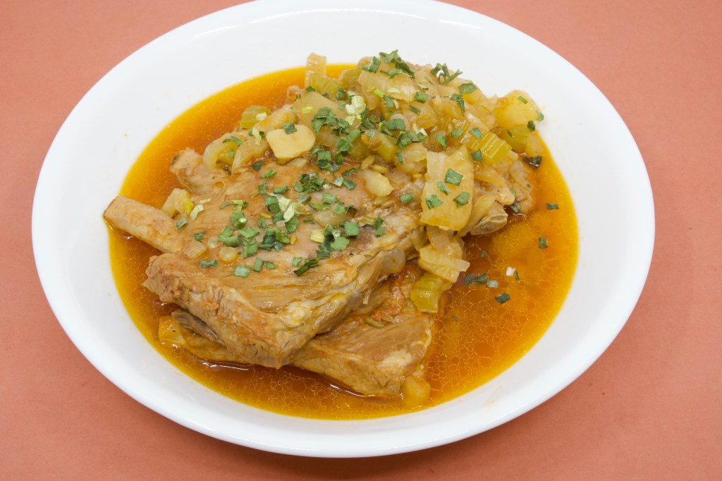 A photograph of a shallow white ceramic bowl containing two racks of pork ribs that had been cooked in the pressure cooker. The ribs are sitting in a thin broth of the cooking liquor, along with soft fennel, onion, and celery. The dish is garnished with some chives.