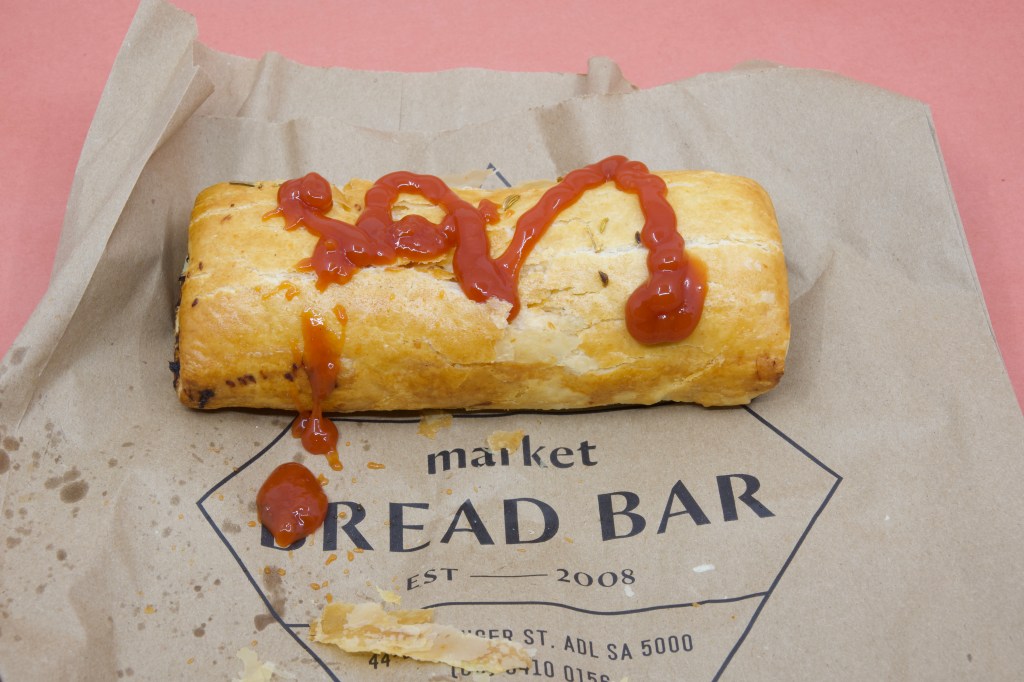 A photograph of a Market Bread Bar Pork and Fennel sausage roll with tomato sauce drizzled on it. The sausage roll is on a Market Bread Bar brown paper bag.