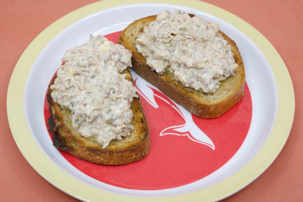 Photograph of John West sardines in spring water with market bread, bar rye sourdough, which has been toasted, and buttered with cultured butter. The sardines have been mixed with some Jalna biodynamic organic yoghurt. The sardines on the two pieces of toasted rye sourdough are placed on a Dolphins NRL plate.