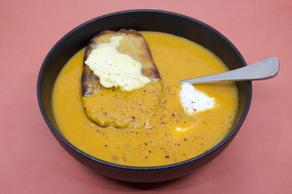 A photograph of pumpkin soup in a black bowl with a large piece of rye sourdough that has cultured butter over it. There is also some yoghurt in the soup that you can see near the spoon. Freshly cracked black pepper is also visible on the soup.