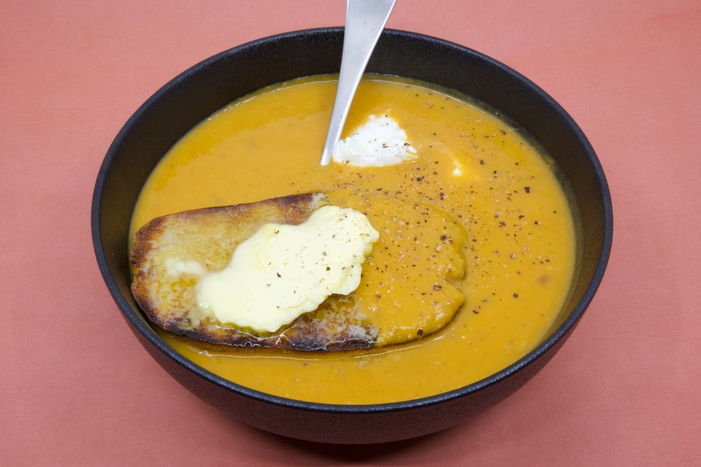 A photograph of pumpkin soup in a black bowl with a large piece of rye sourdough that has cultured butter over it. There is also some yoghurt in the soup that you can see near the spoon. Freshly cracked black pepper is also visible on the soup.