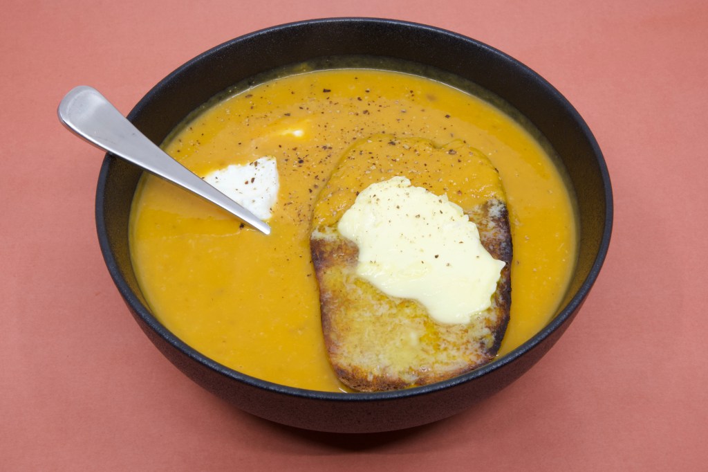 A photograph of pumpkin soup in a black bowl with a large piece of rye sourdough that has cultured butter over it. There is also some yoghurt in the soup that you can see near the spoon. Freshly cracked black pepper is also visible on the soup.
