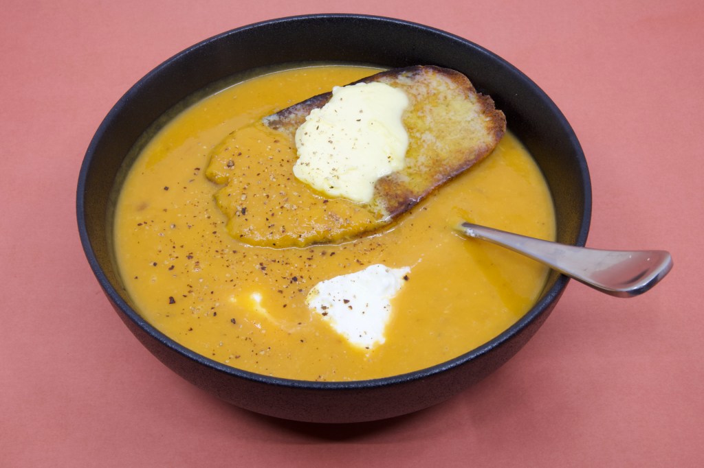 A photograph of pumpkin soup in a black bowl with a large piece of rye sourdough that has cultured butter over it. There is also some yoghurt in the soup that you can see near the spoon. Freshly cracked black pepper is also visible on the soup.