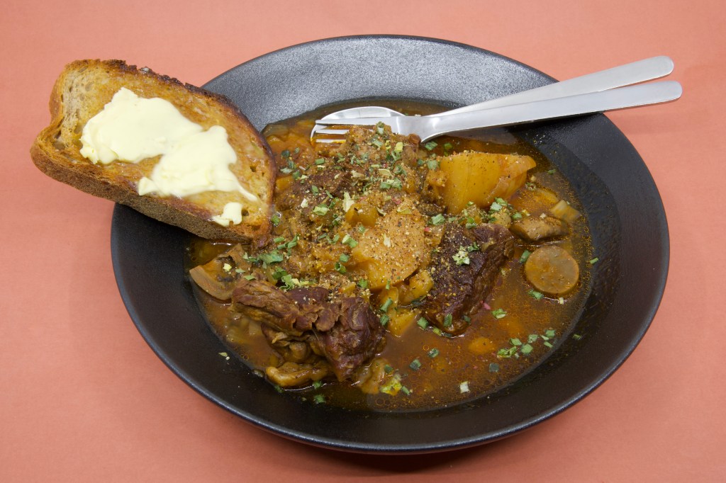 A photograph with a shallow black bowl containing stewed beef chuck and vegetables with a slice of rye sourdough and cultured butter