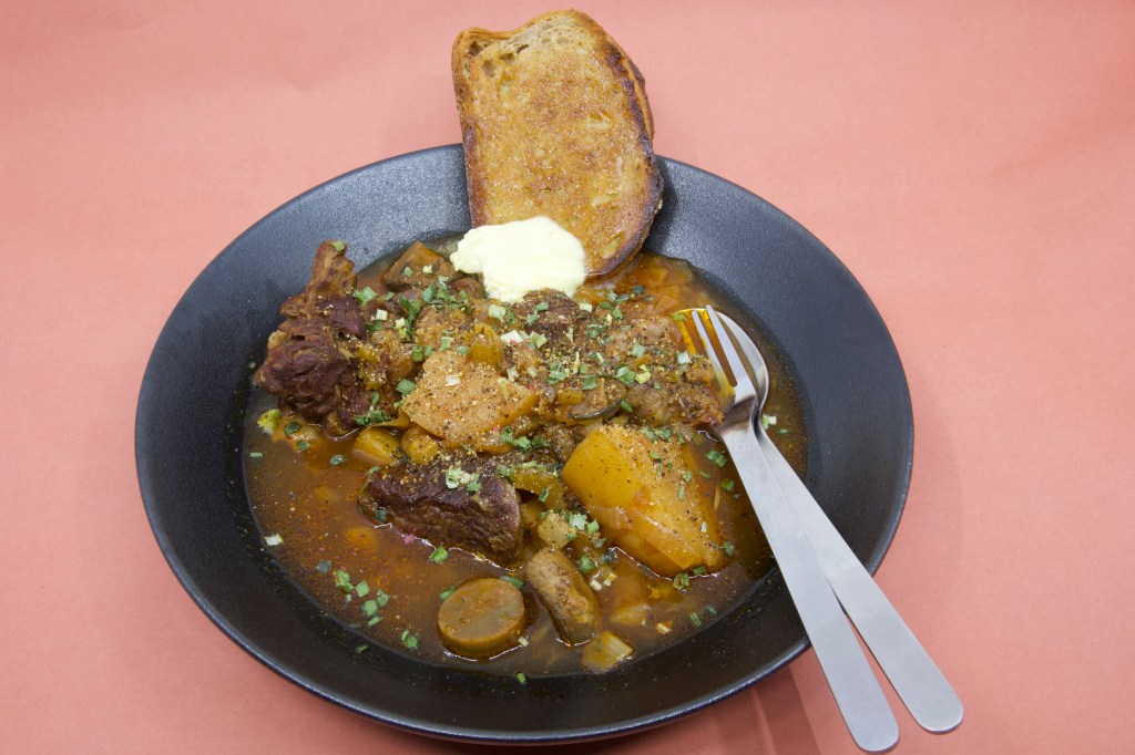 A photograph with a shallow black bowl containing stewed beef chuck and vegetables with a slice of rye sourdough and cultured butter