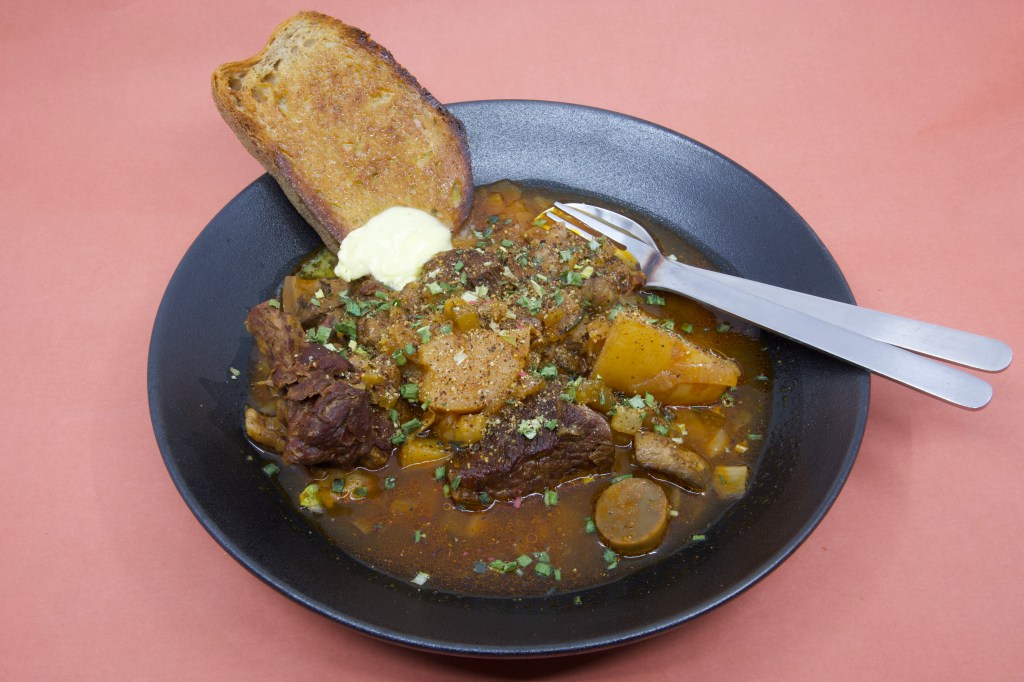 A photograph with a shallow black bowl containing stewed beef chuck and vegetables with a slice of rye sourdough and cultured butter