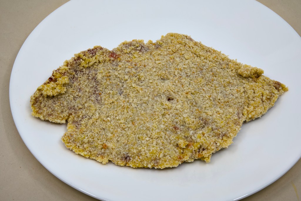 This is a photograph of a white dinner plate with a piece of uncooked crumbed beef on it.