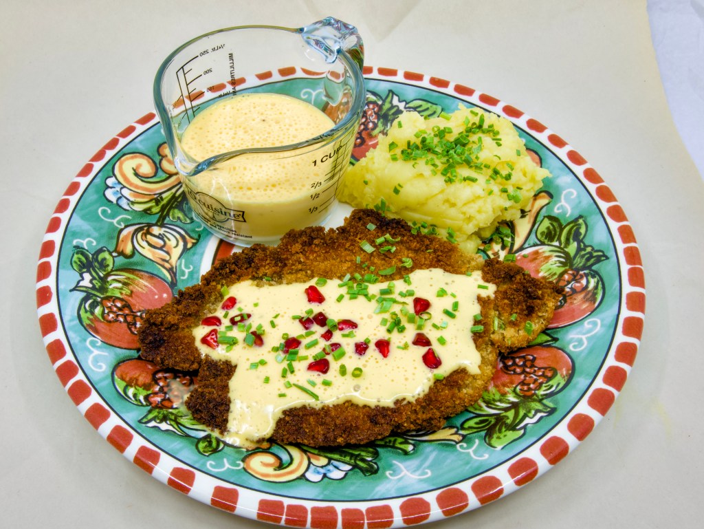 This is a photo of a colourful dinner plate with a beef schnitty (crumbed beef) topped with Hollandaise sauce, served with mashed potato and pickled pomegranate arils. Also known as 'Brother of Courtney.' There is also a small pouring jug of hollandaise sauce on the plate next to the beef schnitty. The mashed potato is garnished with chives.