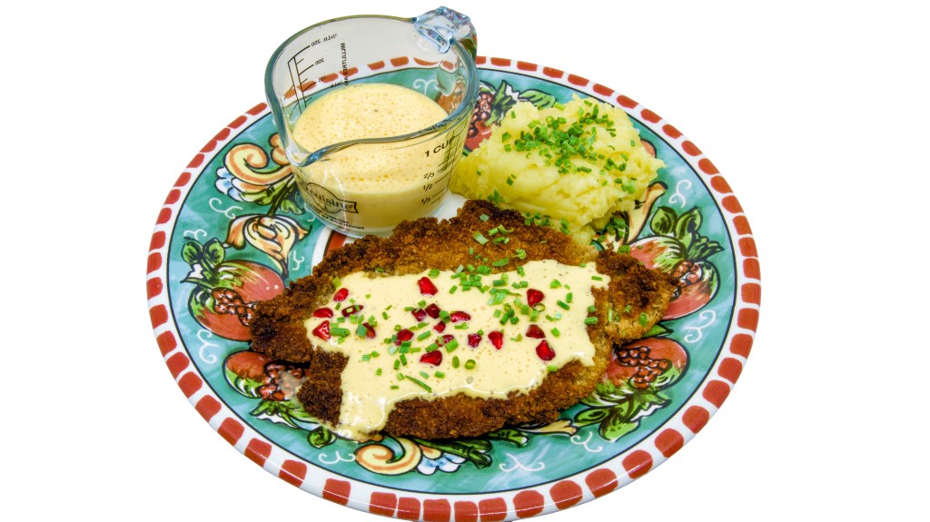 This is a photo of a colourful dinner plate with a beef schnitty (crumbed beef) topped with Hollandaise sauce, served with mashed potato and pickled pomegranate arils. Also known as 'Brother of Courtney.' There is also a small pouring jug of hollandaise sauce on the plate next to the beef schnitty. The mashed potato is garnished with chives.