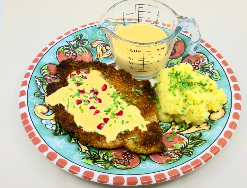 This is a photo of a colourful dinner plate with a beef schnitty (crumbed beef) topped with Hollandaise sauce, served with mashed potato and pickled pomegranate arils. Also known as 'Brother of Courtney.' There is also a small pouring jug of hollandaise sauce on the plate next to the beef schnitty. The mashed potato is garnished with chives.