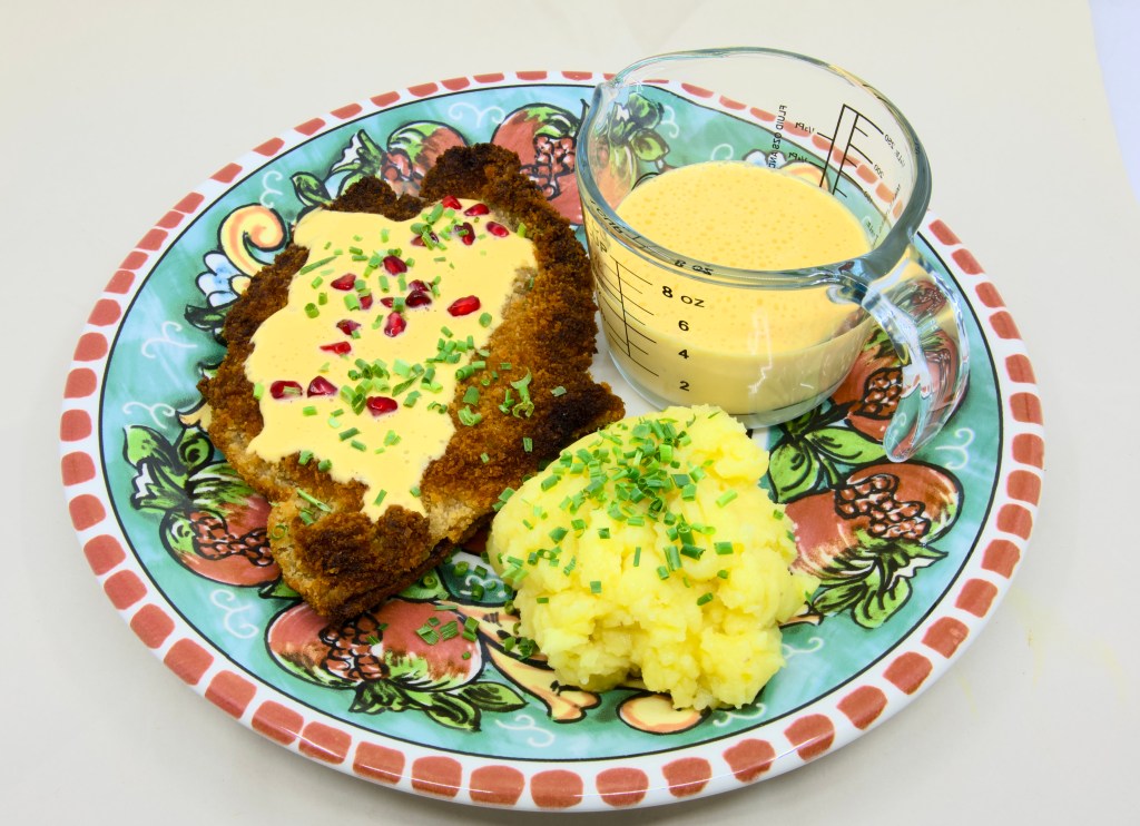 This is a photo of a colourful dinner plate with a beef schnitty (crumbed beef) topped with Hollandaise sauce, served with mashed potato and pickled pomegranate arils. Also known as 'Brother of Courtney.' There is also a small pouring jug of hollandaise sauce on the plate next to the beef schnitty. The mashed potato is garnished with chives.