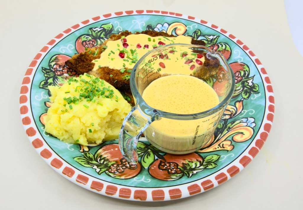 This is a photo of a colourful dinner plate with a beef schnitty (crumbed beef) topped with Hollandaise sauce, served with mashed potato and pickled pomegranate arils. Also known as 'Brother of Courtney.' There is also a small pouring jug of hollandaise sauce on the plate next to the beef schnitty. The mashed potato is garnished with chives.