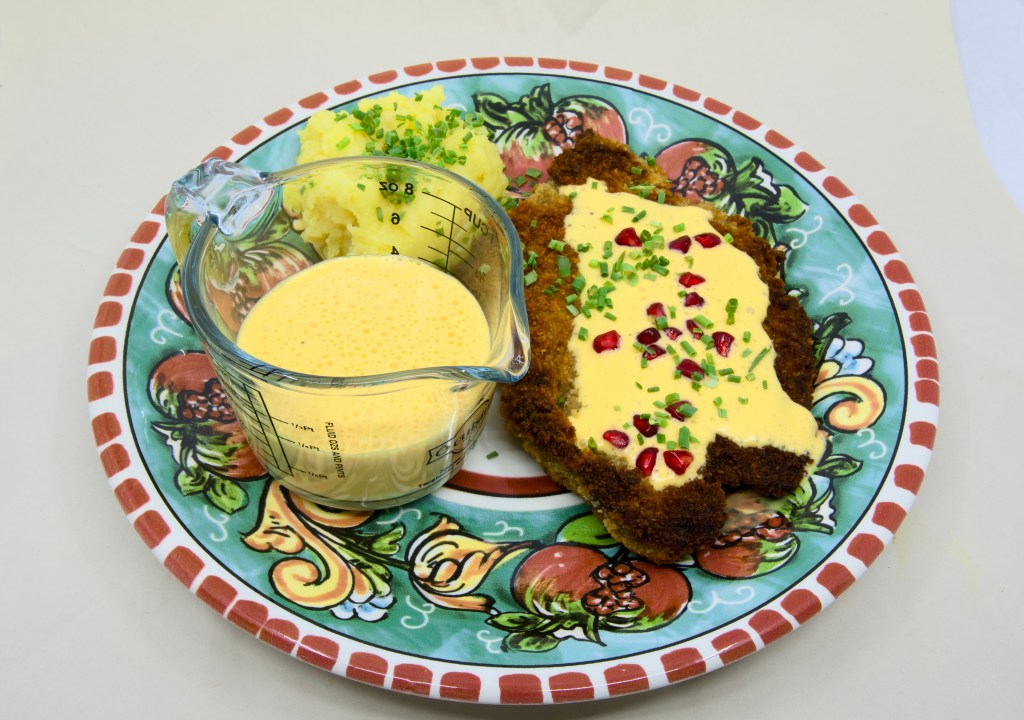 This is a photo of a colourful dinner plate with a beef schnitty (crumbed beef) topped with Hollandaise sauce, served with mashed potato and pickled pomegranate arils. Also known as 'Brother of Courtney.' There is also a small pouring jug of hollandaise sauce on the plate next to the beef schnitty. The mashed potato is garnished with chives.