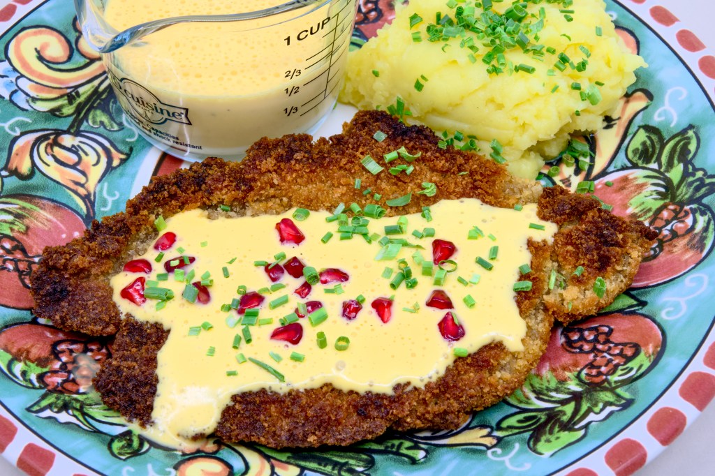 This is a photo of a colourful dinner plate with a beef schnitty (crumbed beef) topped with Hollandaise sauce, served with mashed potato and pickled pomegranate arils. Also known as 'Brother of Courtney.' There is also a small pouring jug of hollandaise sauce on the plate next to the beef schnitty. The mashed potato is garnished with chives.