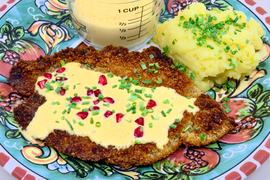 This is a photo of a colourful dinner plate with a beef schnitty (crumbed beef) topped with Hollandaise sauce, served with mashed potato and pickled pomegranate arils. Also known as 'Brother of Courtney.' There is also a small pouring jug of hollandaise sauce on the plate next to the beef schnitty. The mashed potato is garnished with chives.